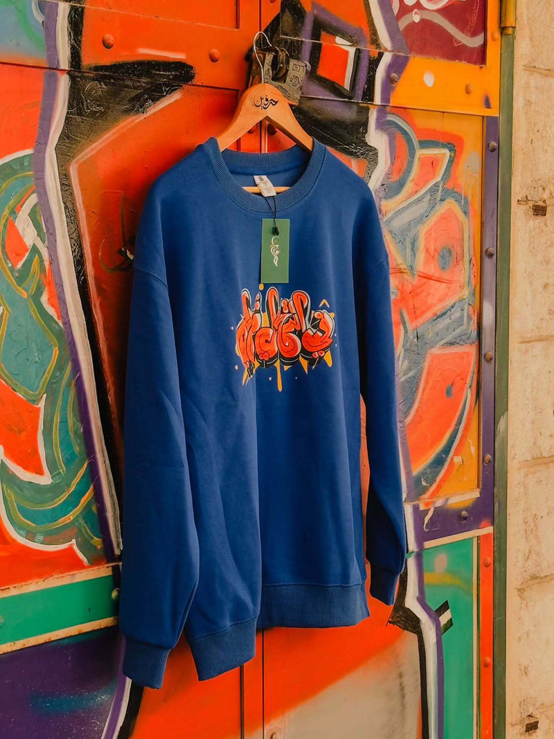 Blue sweatshirt with a colorful graphic design hanging on a hanger against a vibrant, abstract wall.