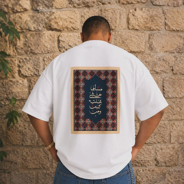 Person wearing a white t-shirt with Arabic text and design against a stone wall.